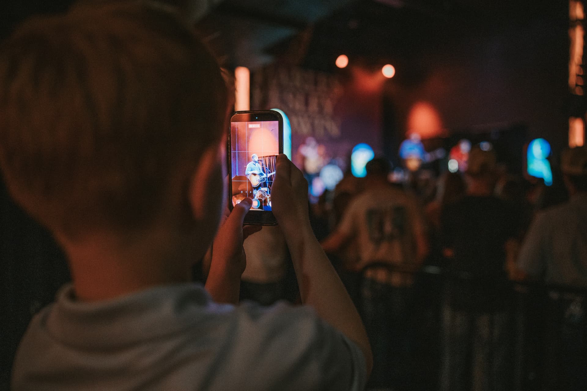 A young fan films an acoustic performance on a phone inside a warmly lit club