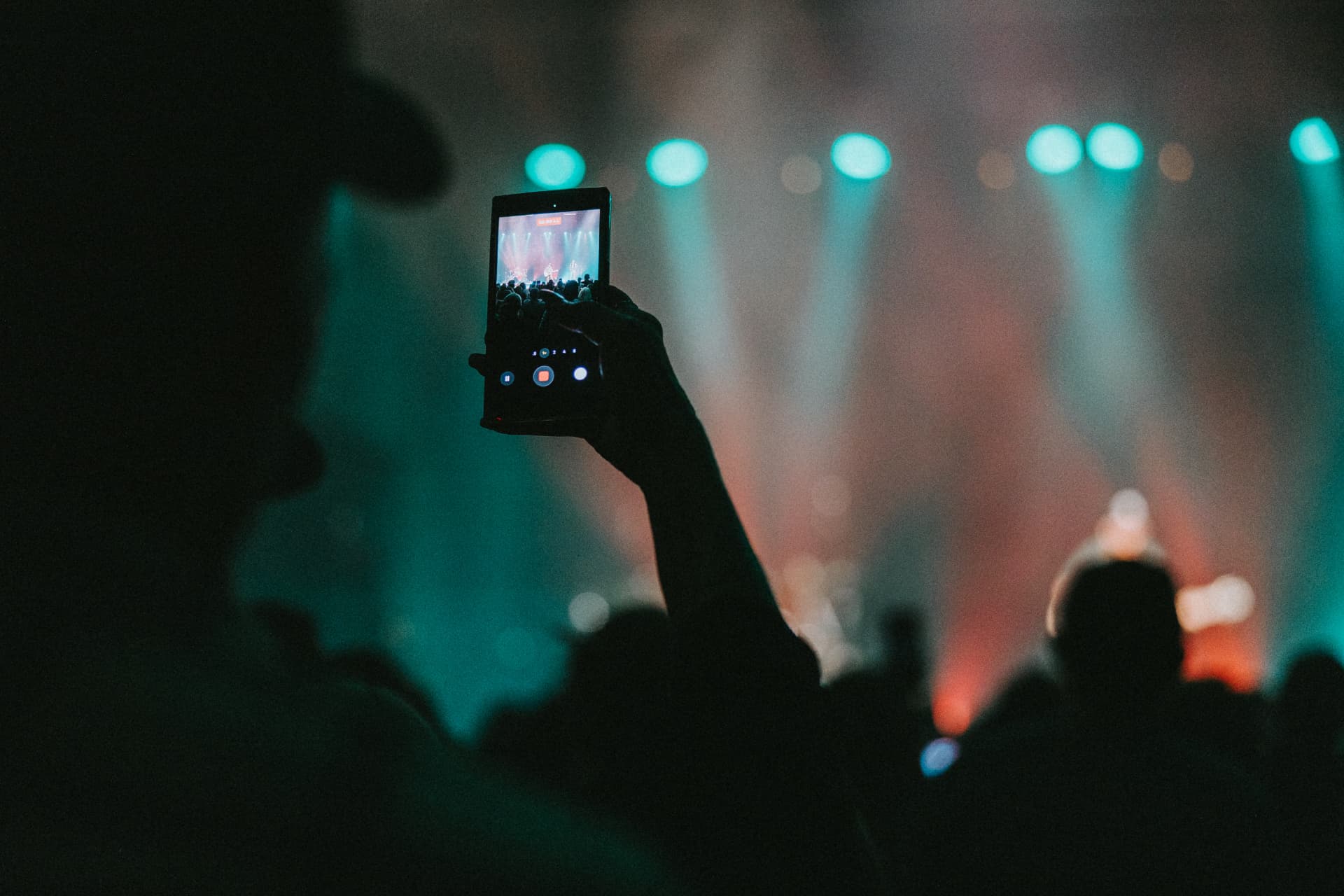Hand raised high holding a phone recording a band under teal stage beams