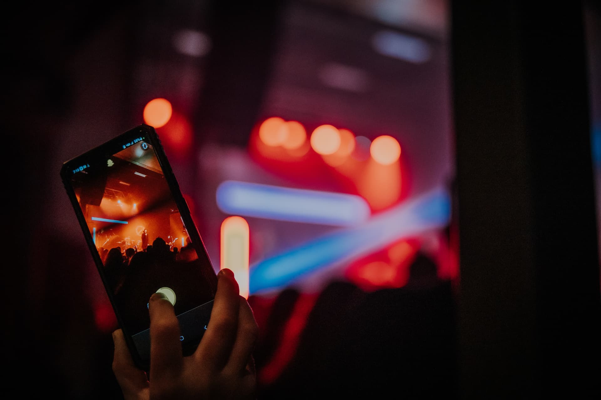 A phone held up in a dark venue recording a performer, framed by red and blue neon lights