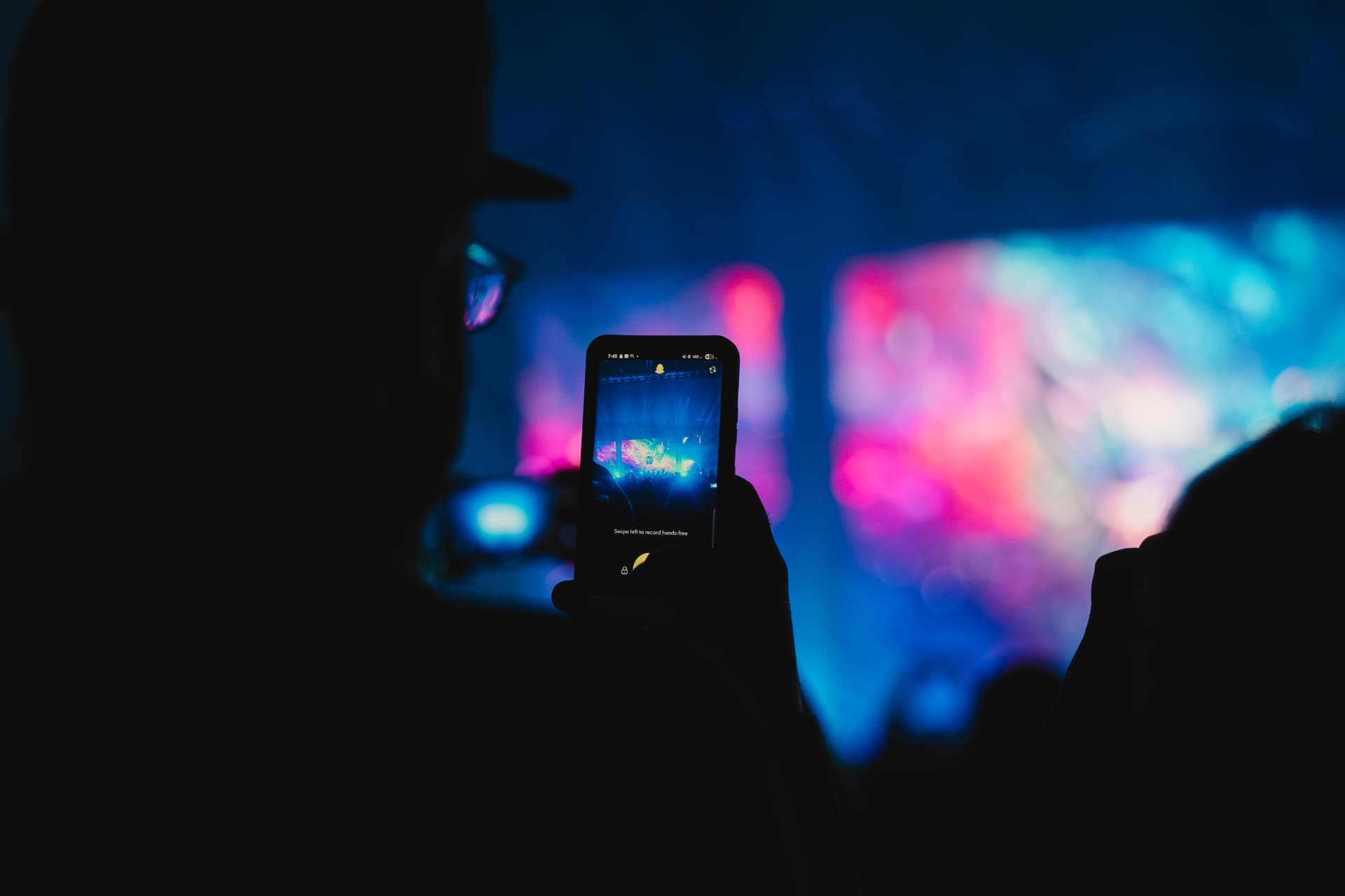 Silhouette of a man filming a concert on his phone as pink and blue stage lights glow behind him