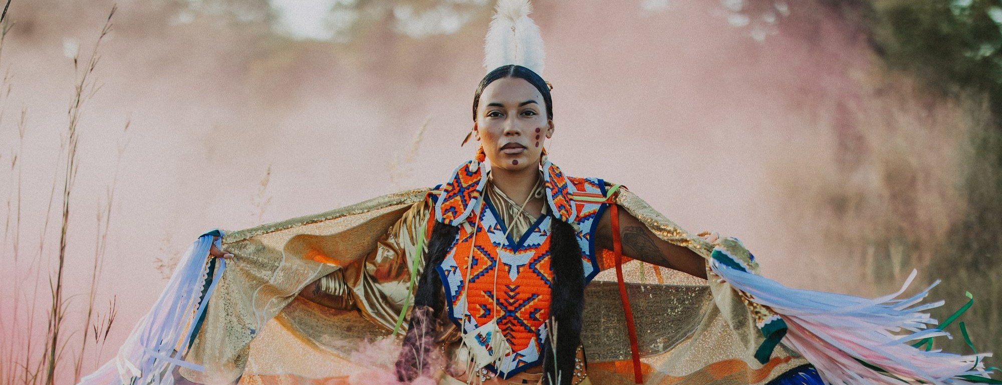 Caitlin Tate Thunder in her fancy shawl regalia, arms outstretched in a prairie field as colored smoke drifts around her