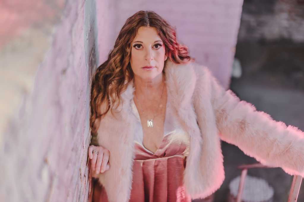 A woman in a pink fur coat leaning against a wall.